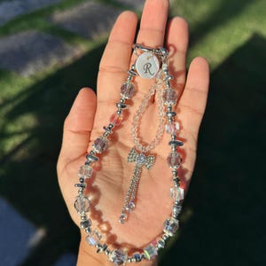 May include: A beaded phone charm and a matching necklace. The phone charm features clear, pink, and silver beads, with a small charm with the letter "R". The necklace has a bow pendant with a dangling chain.