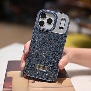 May include: A blue and silver phone case with a sparkling, beaded design. The case has a silver camera frame with three lenses and a single lens, and is embellished with small crystals. A small gold-colored Swarovski logo is visible.