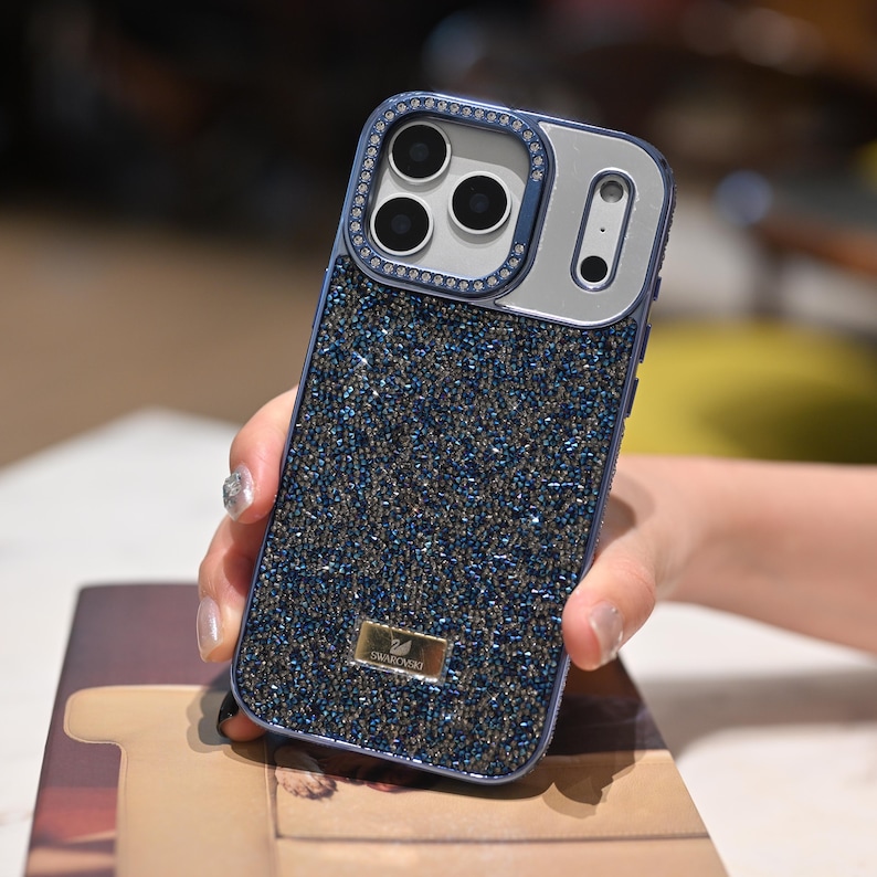 May include: A blue and silver phone case with a sparkling, beaded design. The case has a silver camera frame with three lenses and a single lens, and is embellished with small crystals. A small gold-colored Swarovski logo is visible.