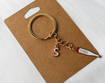 May include: Gold-coloured keyring with a pink letter "S" charm and a white and red thermometer charm. The keyring is attached to a brown cardboard tag.