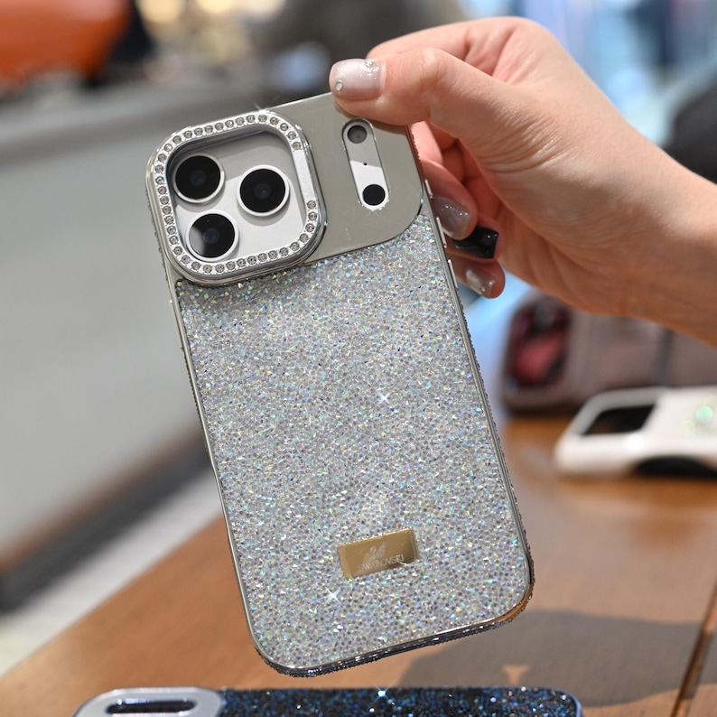 May include: A silver and gray phone case with a sparkling, glittery design. The case has a camera lens protector with a ring of small, clear stones. A small, rectangular gold-colored emblem is on the lower part of the case.