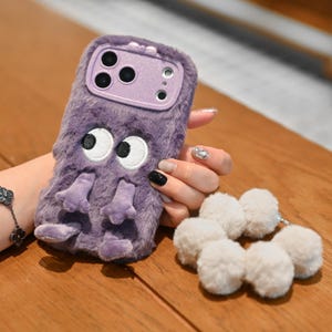 Pode incluir: Uma capa de telefone roxa e difusa com um design de monstro de desenho animado. A capa apresenta olhos brancos grandes, braços e pernas roxos pequenos e uma moldura de câmera roxa combinando. Uma corrente de pompons brancos está presa.