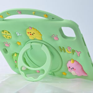 May include: A light green tablet case featuring cartoon dinosaur and star decorations, with the word "HAPPY" on the side. It has a handle and a ring holder. The case is designed to protect a tablet.