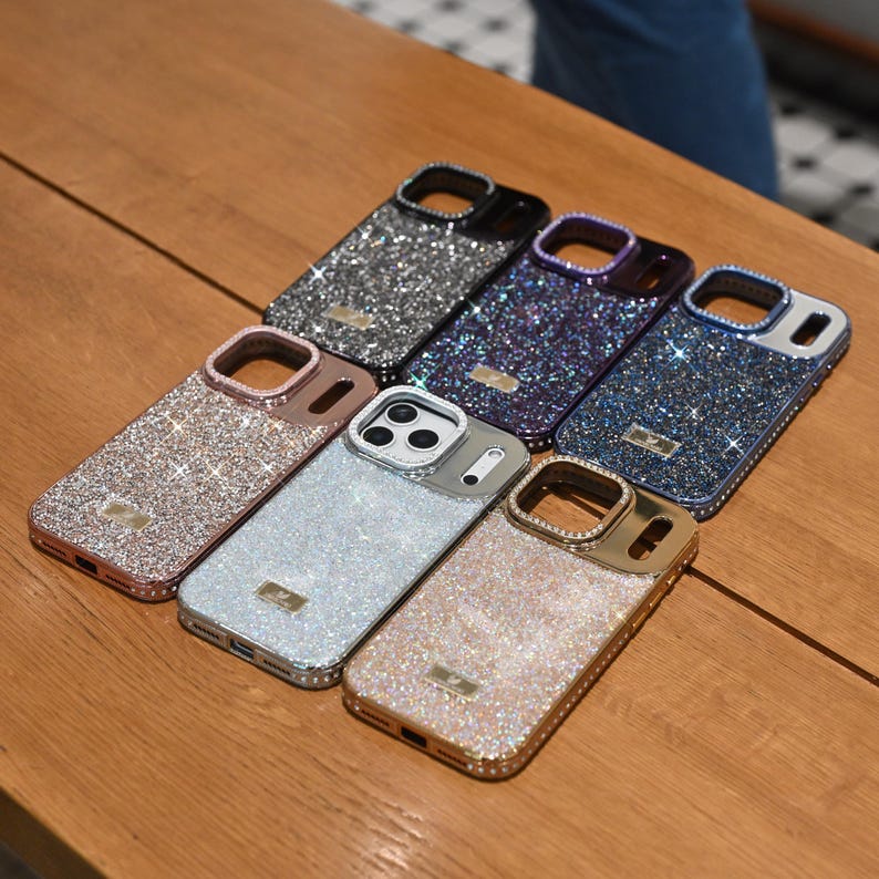 May include: A collection of glittery phone cases in various colors, including silver, pink, gold, and blue, arranged on a wooden surface. Each case has a camera cutout and a small rectangular emblem. The cases are designed to protect the phone while adding a touch of sparkle.