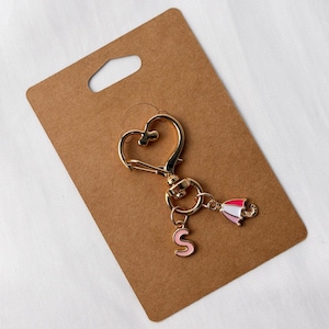 Pode incluir: Chaveiro dourado com fecho em forma de coração, um pequeno pingente com a letra "S" rosa e um pingente de guarda-chuva em miniatura com listras rosa e brancas. Apresentado em um cartão marrom.