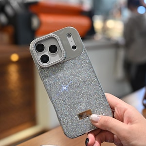 May include: A silver phone case with a sparkling, glittery surface. The camera area is outlined with small, clear crystals. The case has a rectangular shape and a small, rectangular metal tag.