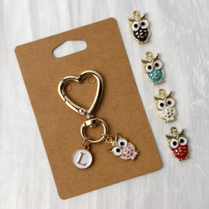 Puede incluir: Un llavero dorado en forma de corazón con un pequeño dije de búho rosa y un dije circular blanco con la letra "L". También hay varios dijes de búho coloridos en negro, verde azulado, blanco y rojo.