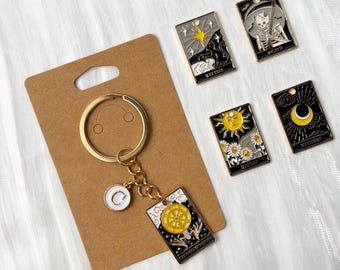 Könnte beinhalten: Goldfarbener Schlüsselanhänger mit einem weißen und goldenen Initialen-Anhänger und einem Tarotkarten-Anhänger. Das Bild zeigt auch vier Tarotkarten-Anhänger mit himmlischen und floralen Designs, darunter The Star, Death, The Sun und The Moon.