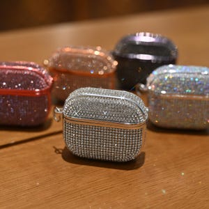 May include: A collection of bedazzled AirPod cases in various colors, including silver, pink, and iridescent. Each case is covered in small, sparkling rhinestones, creating a glamorous and eye-catching accessory. The cases are displayed on a wooden surface.