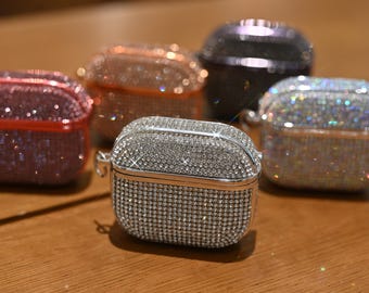 May include: A collection of bedazzled AirPod cases in various colors, including silver, pink, and iridescent. Each case is covered in small, sparkling rhinestones, creating a glamorous and eye-catching accessory. The cases are displayed on a wooden surface.