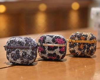 May include: Three bejeweled AirPod cases in a row. The cases are covered in sparkling rhinestones with a leopard print design. The colors include black, silver, pink, and gold. The cases are on a wooden surface.