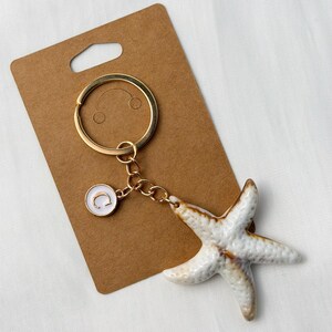 Op de afbeelding: Een goudkleurige sleutelhanger met een witte en bruine zeesterbedel. De sleutelhanger heeft ook een kleine, ronde bedel met een witte achtergrond en de gouden letter "C". De sleutelhanger is bevestigd aan een bruin kartonnen label.
