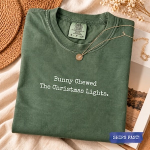 May include: A forest green short-sleeve t-shirt with a crew neckline and the white text "Bunny Chewed The Christmas Lights." printed on the front. The shirt features a Comfort Colors brand label at the inner neck and a gold-toned necklace.