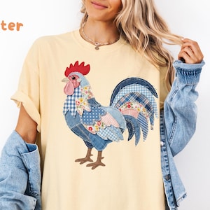 May include: A butter yellow short-sleeve t-shirt featuring a large graphic of a rooster made from various blue, white, and floral patterned fabric patches. The rooster has a red comb and wattle, with brown legs. The word butter is printed in orange.