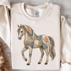 May include: A cream-colored sweatshirt featuring a detailed illustration of a horse. The horse is adorned with a patchwork design in shades of blue, green, and pink, with floral accents. The sweatshirt has long sleeves and a crew neck.