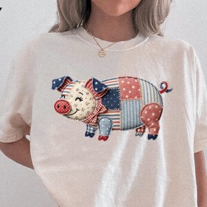 May include: Ivory-colored t-shirt featuring a patchwork pig design in red, white, and blue. The pig has a floral face, a red nose, and a bow tie. The design is reminiscent of the American flag.