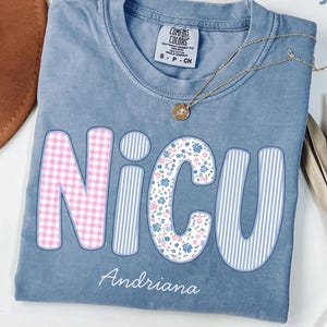 May include: Blue jean Comfort Colors t-shirt with the word "NICU" in a bubble font. The "N" is pink gingham, the "I" is blue and white stripes, the "C" is floral, and the "U" is blue and white stripes. The name "Andriana" is printed below.