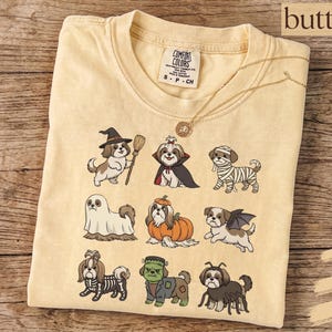 Op de afbeelding: Een boterkleurig t-shirt met een grafische print van negen Shih Tzu-honden in verschillende Halloween-kostuums, waaronder een heks, vampier, mummie, geest, pompoen, vleermuis, skelet, het monster van Frankenstein en een spin.