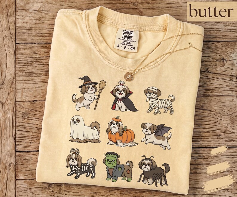 Op de afbeelding: Een boterkleurig t-shirt met een grafische print van negen Shih Tzu-honden in verschillende Halloween-kostuums, waaronder een heks, vampier, mummie, geest, pompoen, vleermuis, skelet, het monster van Frankenstein en een spin.