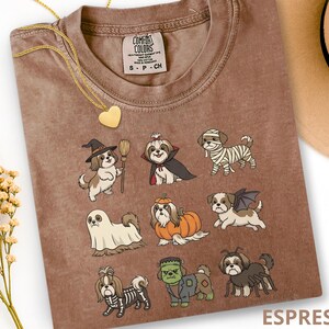 Op de afbeelding: Een bruin t-shirt met een grafische print van negen Shih Tzu honden in verschillende Halloween-kostuums, waaronder een heks, vampier, mummie, spook, pompoen, vleermuis, skelet, het monster van Frankenstein en een spin.