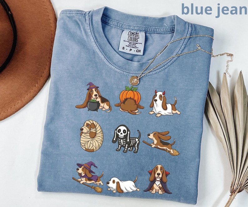 Pu&ograve; includere: Una t-shirt color blu jeans con una stampa grafica di nove cani Basset Hound in vari costumi di Halloween, tra cui una strega, una zucca, un fantasma, una mummia, uno scheletro e un vampiro. La maglietta presenta un'etichetta Comfort Colors.