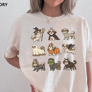 Op de afbeelding: Een ivoorkleurig t-shirt met een grafische print van negen Shih Tzu honden in verschillende Halloween-kostuums, waaronder een heks, vampier, mummie, spook, pompoen, vleermuis, skelet, Frankenstein's monster en een spin, gerangschikt in een raster.
