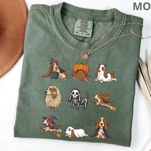 Pu&ograve; includere: Una t-shirt a girocollo color verde muschio con una stampa grafica di nove basset hound in vari costumi di Halloween, tra cui una strega, una zucca, un fantasma, una mummia, uno scheletro e un vampiro. La maglietta ha un'etichetta Comfort Colors.
