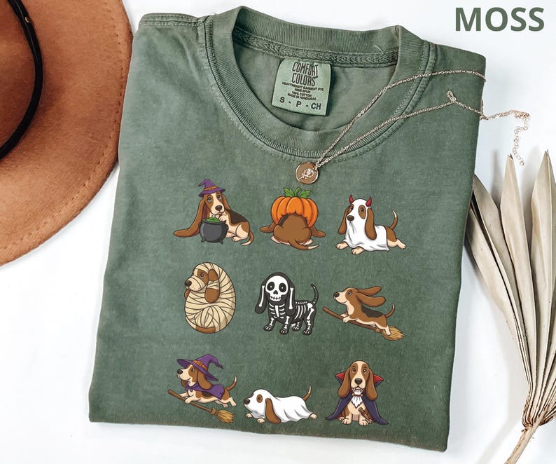 Pu&ograve; includere: Una t-shirt a girocollo color verde muschio con una stampa grafica di nove basset hound in vari costumi di Halloween, tra cui una strega, una zucca, un fantasma, una mummia, uno scheletro e un vampiro. La maglietta ha un'etichetta Comfort Colors.