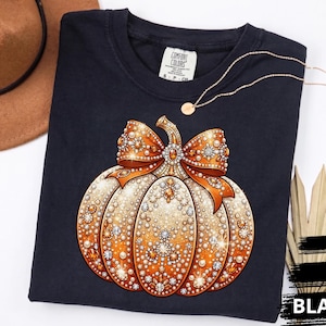 May include: A black short-sleeved t-shirt featuring a graphic of a pumpkin decorated with orange and white rhinestones and a large matching bow. The shirt has a Comfort Colors brand label at the neck and is displayed with a brown hat and dried leaves.