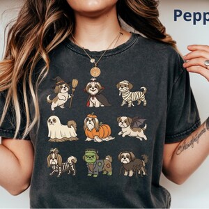 Op de afbeelding: Een antracietgrijs t-shirt met een grafische print van negen Shih Tzu honden in verschillende Halloween-kostuums, waaronder een heks, vampier, mummie, spook, pompoen, vleermuis, skelet, het monster van Frankenstein en een spin.