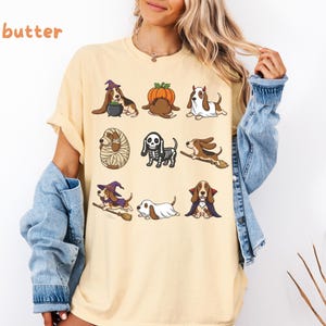 Pu&ograve; includere: Una t-shirt color burro con una stampa grafica di nove cani Basset Hound in vari costumi di Halloween, tra cui una strega, una zucca, un fantasma, una mummia, uno scheletro, un vampiro e cani su manici di scopa, con il testo butter.