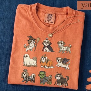 Op de afbeelding: Een oranje T-shirt met korte mouwen en een grafische print van negen Shih Tzu-honden in verschillende Halloween-kostuums, waaronder een heks, vampier, mummie, spook, pompoen, vleermuis, skelet, het monster van Frankenstein en een spin.