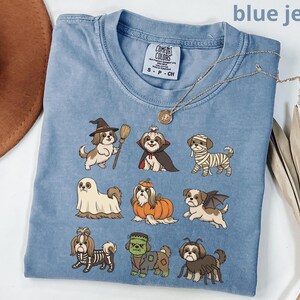 Op de afbeelding: Een blauw spijkerkleurig t-shirt met een grafische print van negen Shih Tzu honden in verschillende Halloween-kostuums, waaronder een heks, vampier, mummie, spook, pompoen, vleermuis, skelet, Frankenstein-monster en een spin.