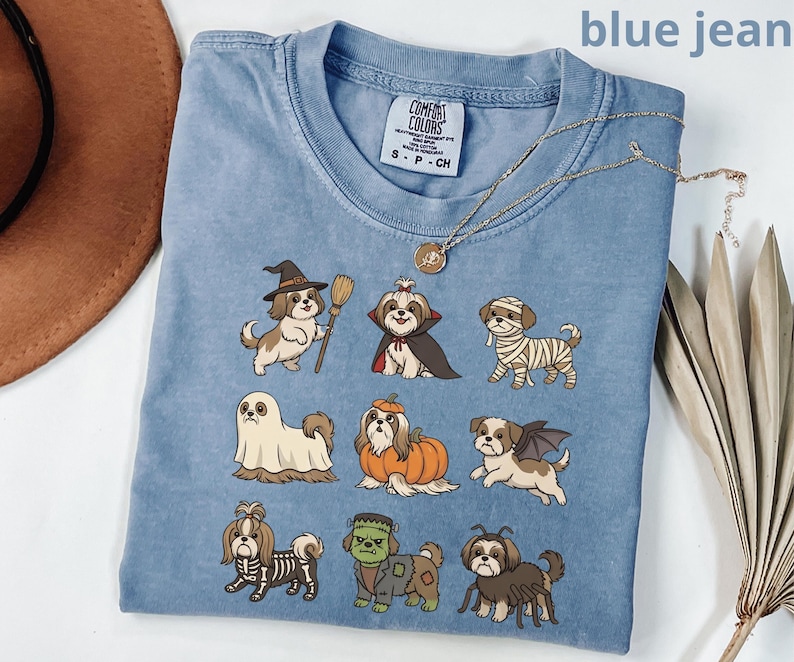 Op de afbeelding: Een blauw spijkerkleurig t-shirt met een grafische print van negen Shih Tzu honden in verschillende Halloween-kostuums, waaronder een heks, vampier, mummie, spook, pompoen, vleermuis, skelet, Frankenstein-monster en een spin.
