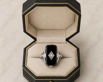 May include: A silver-toned ring featuring a rectangular black onyx stone with a central diamond-shaped inlay holding a small clear gemstone. The ring has intricate metalwork on the sides and is displayed inside an octagonal black jewellery box.