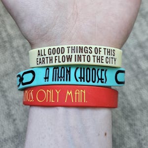 Peut inclure: Trois bracelets en silicone empilés. Le bracelet supérieur est jaune pâle avec le texte noir ALL GOOD THINGS OF THIS EARTH FLOW INTO THE CITY. Le bracelet central est bleu clair avec des maillons de chaîne noirs et le texte A MAN CHOOSES. Le bracelet inférieur est rouge avec le texte jaune GS. ONLY MAN.