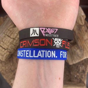 May include: Three rubber wristbands stacked on a wrist. The top band is black with white and pink text and a logo. The middle band is black with red text and a white skull logo. The bottom band is blue with white text.