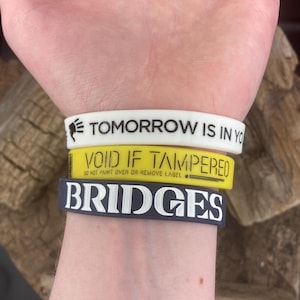 May include: Three silicone wristbands stacked on a person's wrist. The top band is white with black text that reads "TOMORROW IS IN YOU". The middle band is yellow with black text that reads "VOID IF TAMPERED DO NOT PAINT OVER OR REMOVE LABEL". The bottom band is dark gray with white text that reads "BRIDGES".