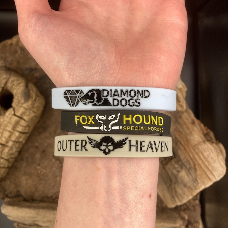 May include: Three silicone wristbands with different designs. The top band is white with black text that reads "Diamond Dogs". The middle band is black with yellow text that reads "Fox Hound - Special Forces". The bottom band is beige with black text that reads "Outer Heaven".