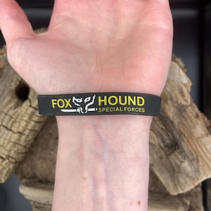 Puede incluir: Pulsera de silicona negra con texto amarillo que dice "FOX HOUND SPECIAL FORCES".