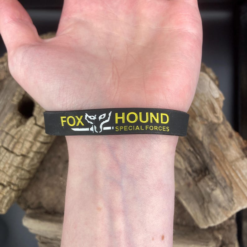 Puede incluir: Pulsera de silicona negra con texto amarillo que dice "FOX HOUND SPECIAL FORCES".
