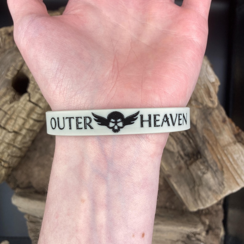 Puede incluir: Una pulsera de silicona blanca con texto negro que dice "OUTER HEAVEN" y una calavera con alas entre las palabras.