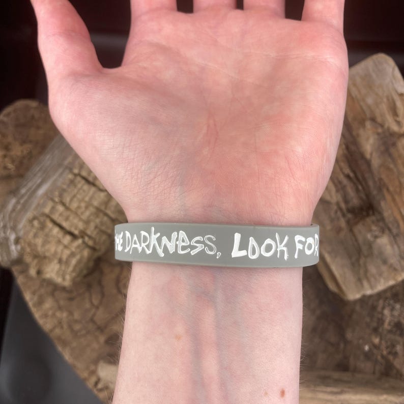 Pu&ograve; includere: Un braccialetto in silicone grigio con il testo "Embrace the darkness, look for the light" in lettere bianche.