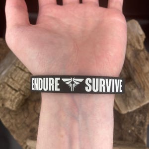 Pu&ograve; includere: Braccialetto in silicone nero con testo bianco che recita "ENDURE" e "SURVIVE" con un simbolo di lucciola tra le parole.