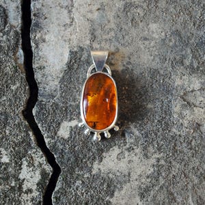 Könnte beinhalten: Silberanhänger mit einem ovalen Bernstein. Der Bernstein hat eine warme, durchscheinende orange-braune Farbe und ist in eine silberne Lünette gefasst. Der Anhänger hat eine kleine Öse oben und ein dekoratives Silberdesign unten.