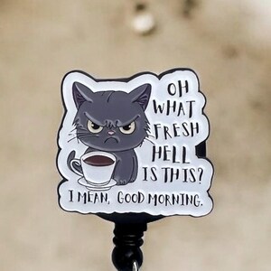 May include: A retractable badge reel with a grumpy gray cat holding a coffee cup. The cat has an angry expression and the text reads "OH WHAT FRESH HELL IS THIS? I MEAN, GOOD MORNING."
