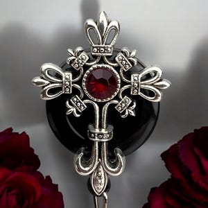 May include: A silver-toned cross pendant with a large, round, red gemstone at its center. The cross features ornate detailing and is set against a black circular backdrop. The design has a gothic or vintage aesthetic.