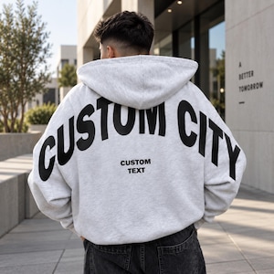 以下が含まれることがあります： 背面に黒い文字でCUSTOM CITYとアーチ状にプリントされ、その下に小さな黒い文字でCUSTOM TEXTと記されたライトグレーのフード付きスウェットシャツ。長袖でフードが付いており、ダークグレーのデニムジーンズの上に着用されています。