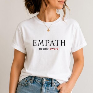 Può includere: T-shirt bianca con la scritta "EMPATH" in nero e "deeply aware" in rosso. La t-shirt è indossata con jeans blu. La persona indossa una collana d'oro.