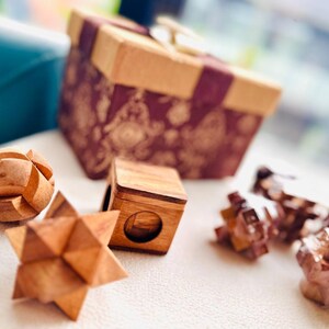 May include: A selection of wooden puzzles and a gift box. The puzzles include a star, a cube, and a ball. The gift box is brown with a patterned design and a ribbon.